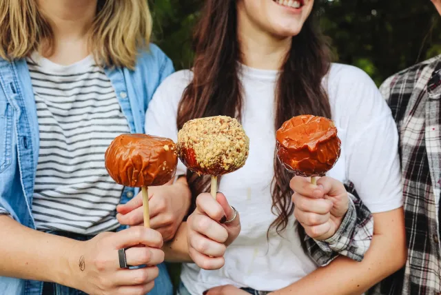 Three friends hold caramel-coated apples, one with nuts, smiling outdoors, showcasing a fun, seasonal treat.