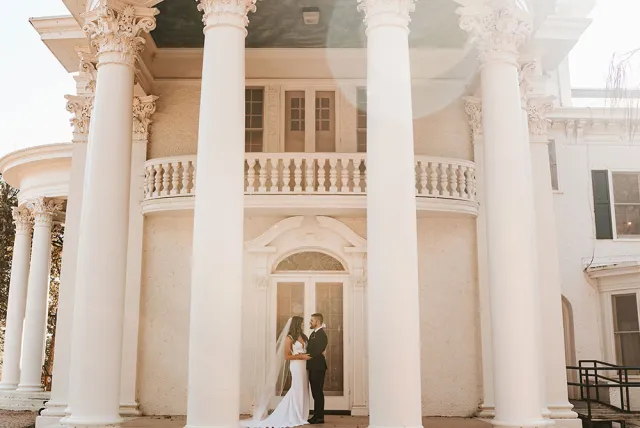 A bride and groom share a tender moment in front of an elegant, columned mansion on their wedding day.