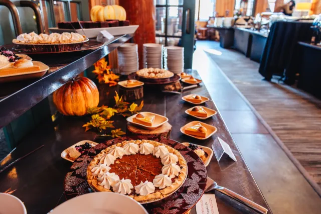 Festive dessert buffet with pumpkin pies topped with whipped cream, surrounded by plates of cheesecake, autumn leaves, and a decorative pumpkin.