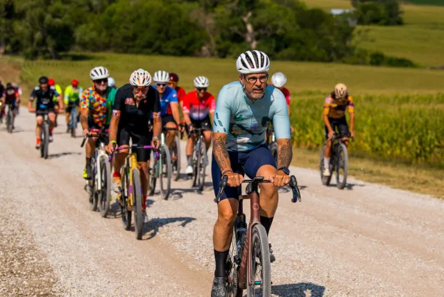 Group of cyclists riding on a gravel road through a rural landscape with green fields and trees on a sunny day.