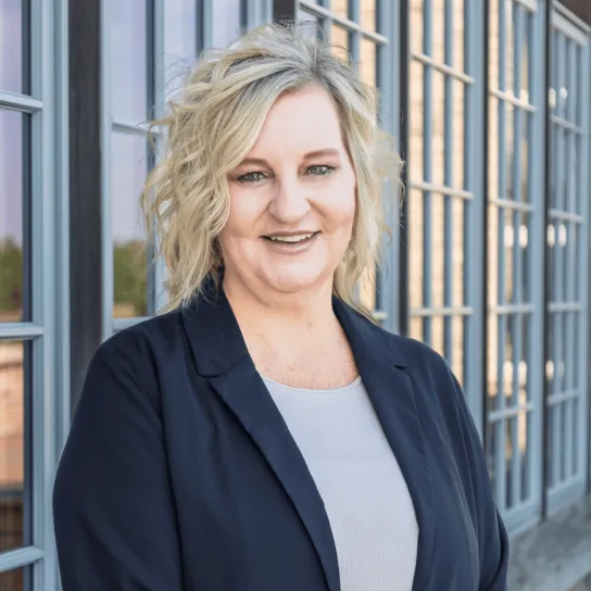 Professional portrait of a woman in a black blazer smiling and standing outside with a windowed stone building in the background.