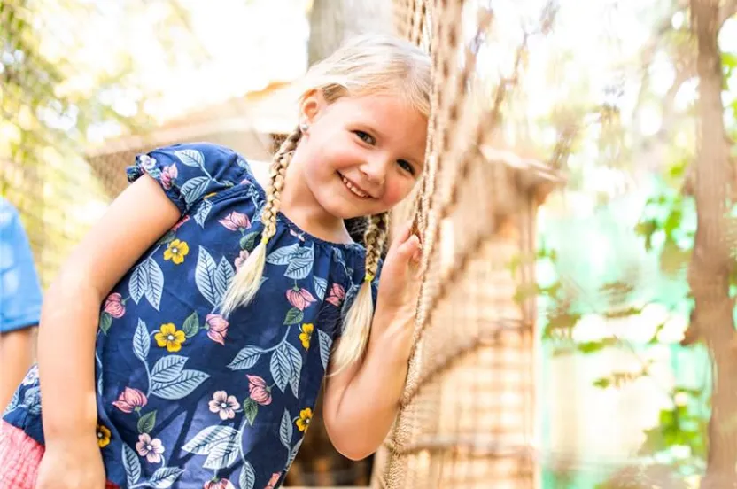 A little girl smiles brightly while standing confidently on a rope bridge, surrounded by greenery.