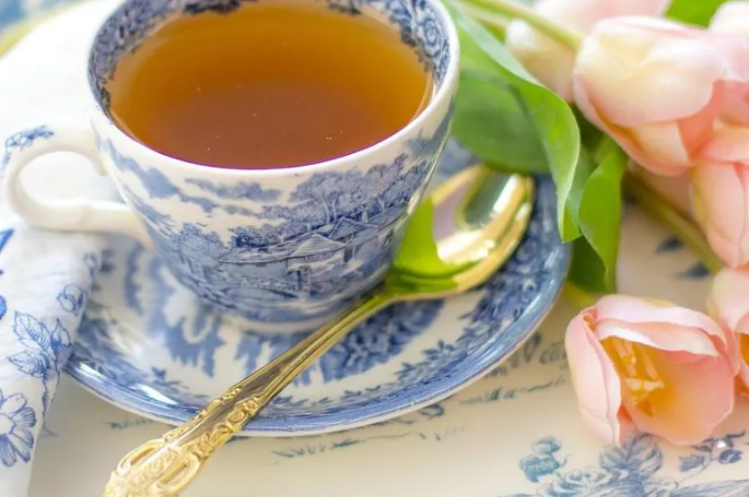 A blue and white porcelain teacup filled with tea, set on an ornate plate alongside a gold spoon and pink tulips.