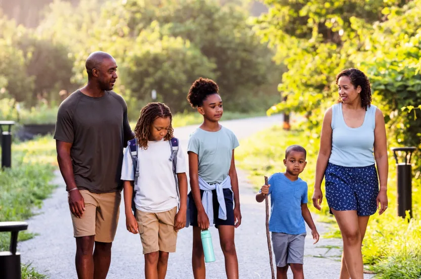 A family of five strolls along a scenic gravel path surrounded by lush greenery, enjoying a sunny day outdoors.