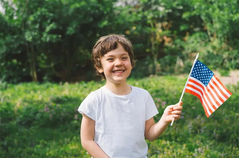 A child in a white t-shirt stands outdoors, holding a small American flag with greenery in the background.