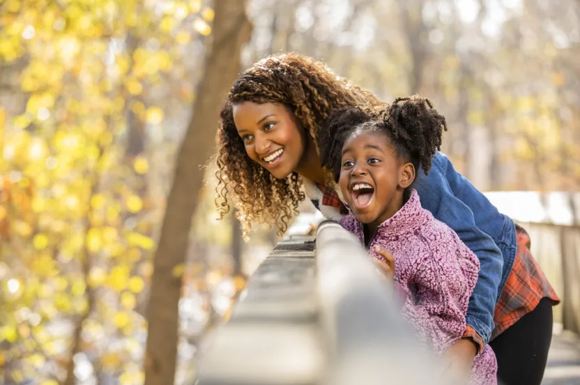 A woman and a child lean over a wooden railing, gazing at a scenic view amid a backdrop of autumn trees and soft, blurred colors.