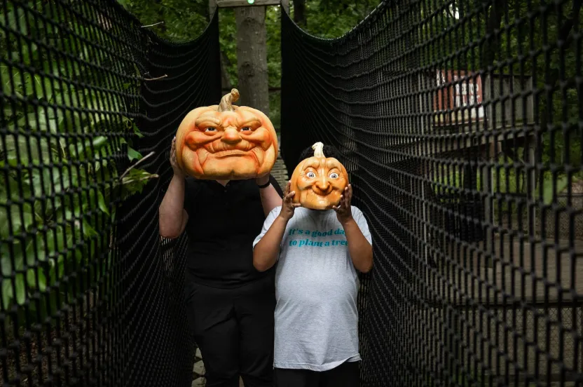 Two individuals standing in a netted pathway, each holding a pumpkin, surrounded by lush greenery.