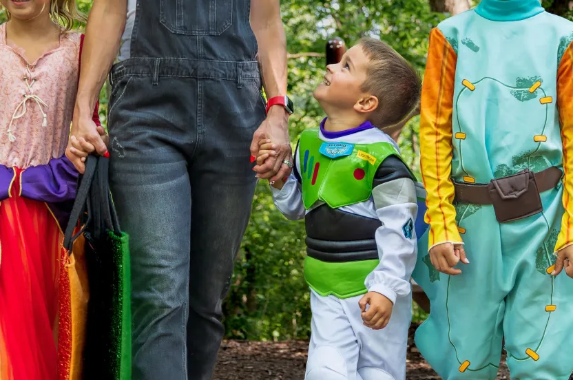 A group of children dressed in colorful costumes, holding hands while walking in a forested area, with an adult companion.
