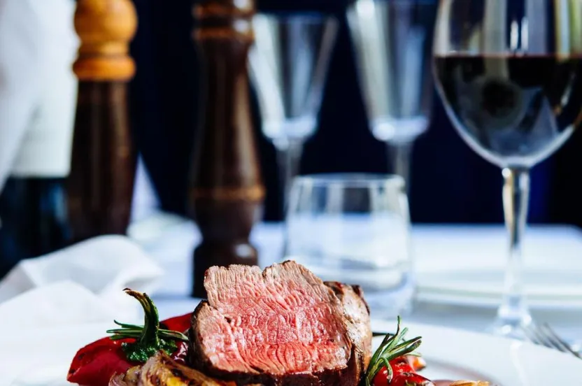 A gourmet dish of medium-rare steak garnished with grilled vegetables on a white plate. A glass of red wine and pepper mills create an elegant dining setting.