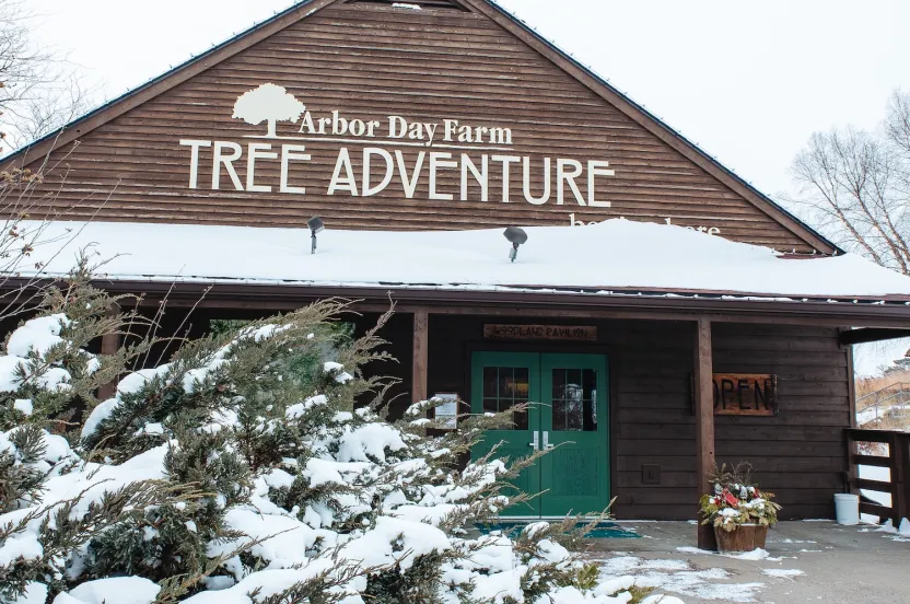 A wooden building with a sign reading "Arbor Day Farm Tree Adventure," surrounded by snow-covered bushes in winter. The doors are green and open.