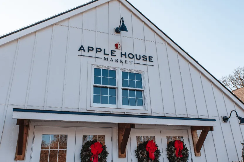The exterior of Apple House Market features a white facade, two windows, holiday wreaths with red bows on the doors, and a sign above the entrance.