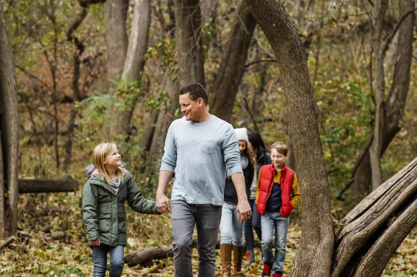 A family walks hand-in-hand through a forested area covered in fallen leaves, surrounded by trees in autumn colors.