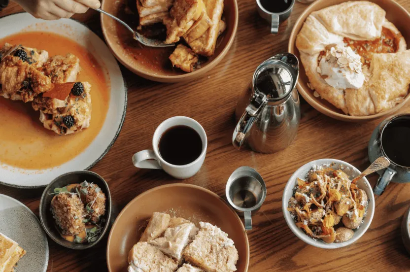 Wooden table with a wide variety of brunch food items and people using forks to eat.