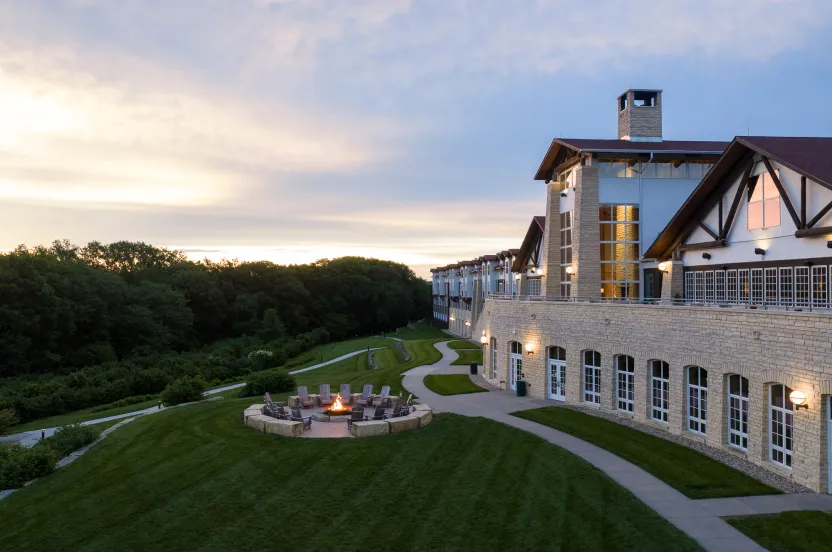 Large stone building with outdoor fire pit surrounded by chairs on a grassy lawn at sunset near a wooded area.