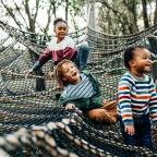 Three children play joyfully in a large net suspended between trees in a wooded area.