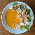 A vibrant orange pannacotta topped with crumbled cookies, herbs, and edible flowers in a white bowl on a wooden table.