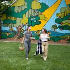 Two women walk on green grass, carrying a basket and a plate, with a colorful mural of trees and landscape behind them.