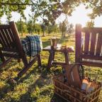 Two rustic wooden chairs with a plaid blanket, a table set with wine and snacks, and a picnic basket, all under a tree at sunset.