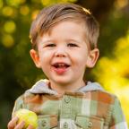 A young child in a plaid jacket holds a green apple, standing outdoors with a blurred green and yellow background.