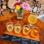A tasting flight of four light drinks labeled 'Russet', 'Joy', 'Tropical Sage', and 'Orange' beside a floral vase on a wooden table.