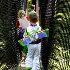 A child in a Buzz Lightyear costume walks on a forest bridge, followed by two other kids, surrounded by lush greenery.