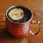 Copper mug with iced cocktail garnished with a dried apple slice on a wooden surface.